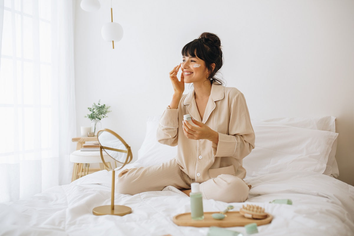 Smiling woman applying face cream sitting on bed