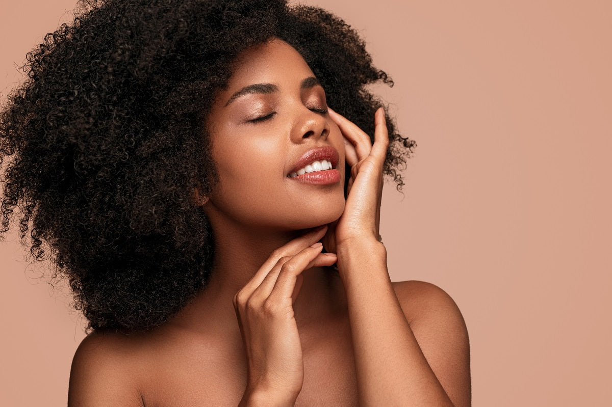 Young female with curly hair enjoying softness of clean face with closed eyes after skin care procedure against brown background